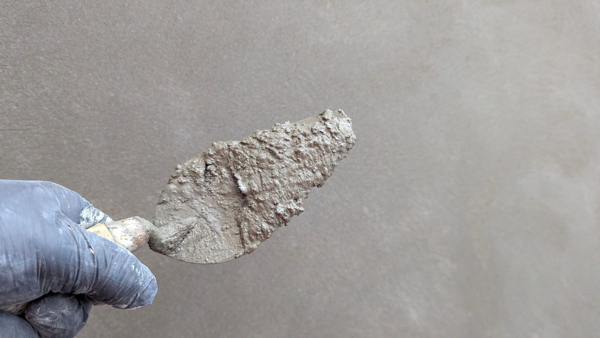 Une main gantée tient une spatule chargée de mortier sur un mur